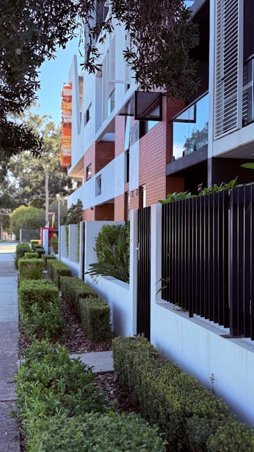 The exterior of a modern residential building on Sussex Gardens, Paddington, features a well-maintained garden with neatly trimmed green hedges and small shrubs along a paved pathway. The building's facade includes a combination of brick and glass materials, with large windows and balconies. The black metal fence with vertical slats runs parallel to the sidewalk, providing a decorative boundary. Natural daylight illuminates the scene, highlighting the clean and orderly appearance of the surrounding outdoor space. As part of their professional cleaning services, Cleaners Paddington ensures that exterior surfaces such as pathways, fences, and building facades remain free of dust, dirt, and grime, maintaining the property's overall appearance and hygiene.
