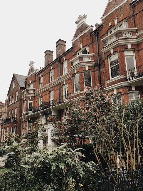Exterior view of a multi-story Victorian-style residential building on Sussex Gardens, Paddington, with red brick facades, decorative white trims, and bay windows. The building features black wrought iron balconies and chimneys on the roof. In the foreground, a garden area with lush green shrubs, flowering plants, and small trees is visible. The scene is captured during daytime with natural light illuminating the building. Cleaners Paddington specializes in domestic cleaning services, including surface cleaning, deep cleaning, and sanitisation, ensuring residential properties like this are maintained in pristine condition.