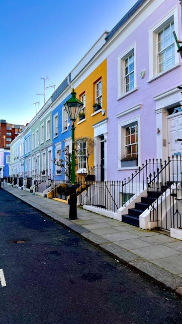 A row of colorful Victorian-style terraced houses in Paddington, featuring facades painted in pastel yellow, pale purple, and light blue. Each house has white-framed sash windows, some with flower boxes on the window sills, and small front steps leading up to white wooden doors. Black wrought iron fences and railings border the front gardens, with a black street lamp positioned on the sidewalk. The pavement appears clean and well-maintained, illuminated by natural daylight under a clear blue sky. This vibrant street scene exemplifies residential character, with no visible cleaning activity or tools present, focusing on the aesthetic appeal of the neighborhood as part of the context for reliable domestic cleaning services by Cleaners Paddington.