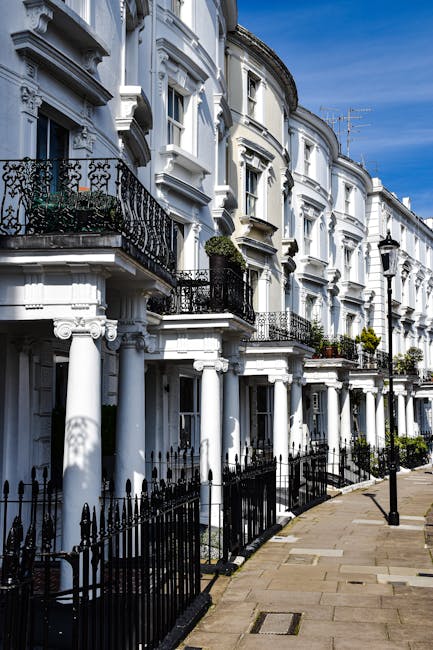 A row of white Victorian-style terraced houses with ornate architectural details, including decorative cornices and intricate iron balcony railings. The buildings feature large sash windows, some with flower boxes and greenery on the balconies. The pavement is clean and well-maintained with black wrought iron fencing along the front of each property. Bright natural sunlight illuminates the scene, highlighting the pristine, freshly cleaned exterior surfaces, which appear smooth and polished. Cleaners Paddington specializes in surface cleaning and deep cleaning, ensuring homes are hygienic and well-maintained as part of their domestic cleaning and sanitisation services. This image emphasizes the importance of regular exterior cleaning to preserve the aesthetic appeal of residential properties.