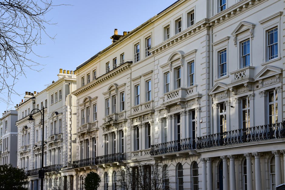 A row of colorful Victorian-style terraced houses in Paddington, featuring facades painted in pastel yellow, pale purple, and light blue. Each house has white-framed sash windows, some with flower boxes on the window sills, and small front steps leading up to white wooden doors. Black wrought iron fences and railings border the front gardens, with a black street lamp positioned on the sidewalk. The pavement appears clean and well-maintained, illuminated by natural daylight under a clear blue sky. This vibrant street scene exemplifies residential character, with no visible cleaning activity or tools present, focusing on the aesthetic appeal of the neighborhood as part of the context for reliable domestic cleaning services by Cleaners Paddington.