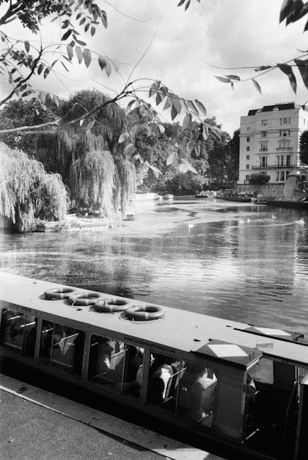 A black and white photograph of a riverside scene in Paddington, showing a glass-paneled boat docked along the edge of the water. The boat has a smooth, reflective surface with visible windows, and appears to be well-maintained. In the background, a large building with multiple floors and balconies is partially obscured by leafy trees, including a prominent weeping willow with long, drooping branches hanging over the river. The river surface is calm, showing gentle ripples, with surrounding vegetation and urban structures creating a tranquil yet structured environment. This image captures the natural and architectural elements typical of Sheldon Square and Merchant Square in Paddington, emphasizing the area’s scenic, clean, and well-kept appearance. Cleaners Paddington would recommend routine surface cleaning and maintenance to preserve the scenic waterways and surrounding elements.