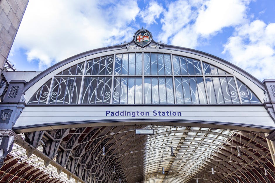 A detailed view of the exterior of Paddington Station, showcasing a large arched glass and steel roof structure with intricate metalwork and reflective glass panels. The sky with some clouds is visible through the glass, creating a bright and airy atmosphere. The station's white and grey color palette contrasts with the blue 'Paddington Station' sign displayed prominently below the arch. Cleaners Paddington specializes in surface cleaning and sanitisation of such architectural exteriors, ensuring high standards of hygiene and visual appeal for both residential and commercial properties in the Paddington area.