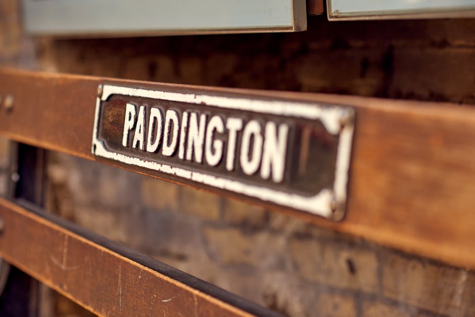 A professional cleaner from Cleaners Paddington, wearing a beige uniform with a name badge, is seen carefully making a bed with white linens in a well-lit, minimalistic room. The room has a plain whit