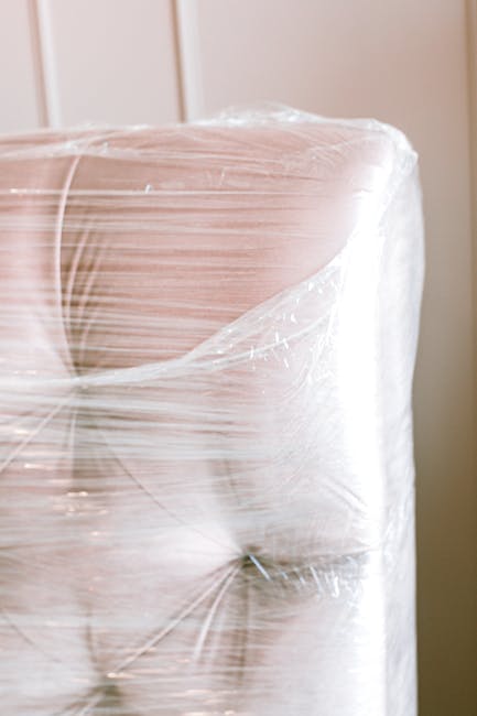Close-up of a large piece of furniture, likely a sofa, wrapped in transparent plastic film for protection during cleaning or storage. The sofa has a soft, light-colored fabric surface, with a visible armrest and cushions. The surface appears clean and well-maintained with no visible dust or stains. The background includes vertical wooden panels and warm indoor lighting, suggesting a residential or commercial setting. The image emphasizes surface protection, which is part of the process of deep cleaning or sanitisation, consistent with services offered by Cleaners Paddington for removing sofa mould and maintaining hygiene. The plastic wrapping indicates preparation for cleaning or post-cleaning protection, aligning with professional cleaning procedures.