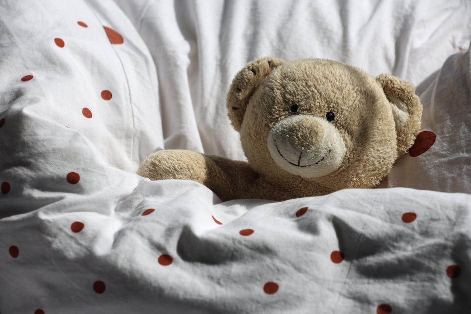 A close-up of a bed with white bedding covered in red polka dots, featuring a plush beige teddy bear with a smiling face placed among the pillows, which are slightly wrinkled. The bed is situated in a well-lit room, with soft natural light highlighting the clean and tidy appearance of the bedding. The background includes additional pillows and a bedding surface, indicating a cozy and hygienic environment. This setting showcases the importance of surface cleaning and maintenance of bedding, aligning with domestic cleaning services provided by Cleaners Paddington, especially for same-day mattress steaming to remove stains and ensure sanitisation in residential spaces.