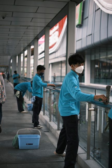 In this indoor commercial setting, several individuals wearing blue uniforms and protective face masks are engaged in surface cleaning along a glass railing and window ledges. They are using cleaning cloths and spray bottles to wipe down the metal handrails and glass surfaces, ensuring thorough sanitisation. The space features large floor-to-ceiling windows allowing natural light to illuminate the area, which includes a tiled floor and an adjacent corridor with a modern ceiling design. A blue cleaning bucket is positioned on the floor nearby, indicating active cleaning procedures. This scene exemplifies diligent hygiene and surface cleaning practices, consistent with the professional standards associated with [COMPANY_NAME] and aligned with the focus on commercial cleaning licences and health and safety duties in Paddington.