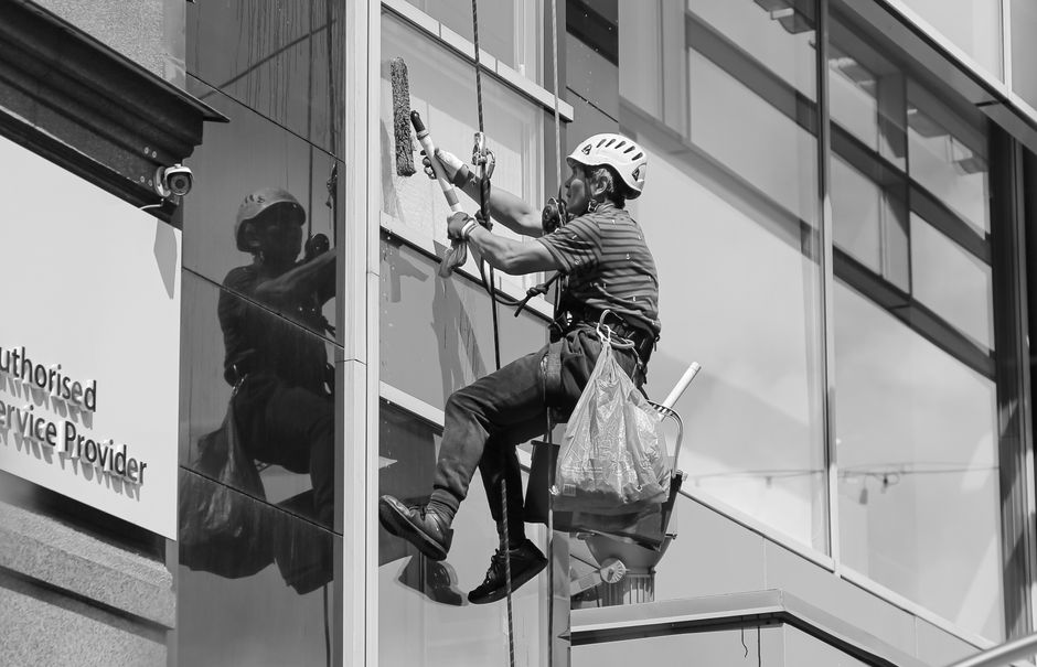 A professional window cleaner wearing safety gear, including a helmet and harness, is cleaning the exterior glass windows of a modern commercial building. The worker is seated in a scaffold basket, holding a squeegee and cloth as they thoroughly wipe the large, reflective glass panels, which are framed by metal mullions. The building surface features a combination of smooth glass and metal cladding, with visible reflections of nearby structures and the sky. The scene is captured during daylight, with natural light illuminating the pristine, spotless windows, highlighting the high standard of surface cleaning and maintenance provided by Cleaners Paddington. Visible tools such as a spray bottle and cleaning cloth are held in the worker's hand, emphasizing the thoroughness of the deep cleaning process involved. This meticulous approach to sanitisation and window surface cleaning exemplifies the professional standards associated with [PAGE_TITLE], ensuring commercial environments are hygienic and presentable.