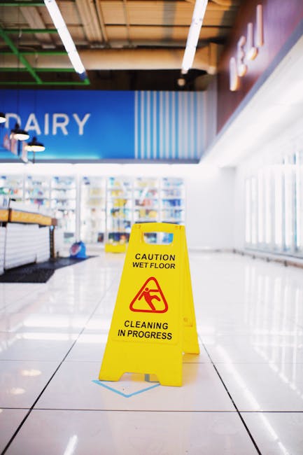In this indoor commercial setting, several individuals wearing blue uniforms and protective face masks are engaged in surface cleaning along a glass railing and window ledges. They are using cleaning cloths and spray bottles to wipe down the metal handrails and glass surfaces, ensuring thorough sanitisation. The space features large floor-to-ceiling windows allowing natural light to illuminate the area, which includes a tiled floor and an adjacent corridor with a modern ceiling design. A blue cleaning bucket is positioned on the floor nearby, indicating active cleaning procedures. This scene exemplifies diligent hygiene and surface cleaning practices, consistent with the professional standards associated with [COMPANY_NAME] and aligned with the focus on commercial cleaning licences and health and safety duties in Paddington.